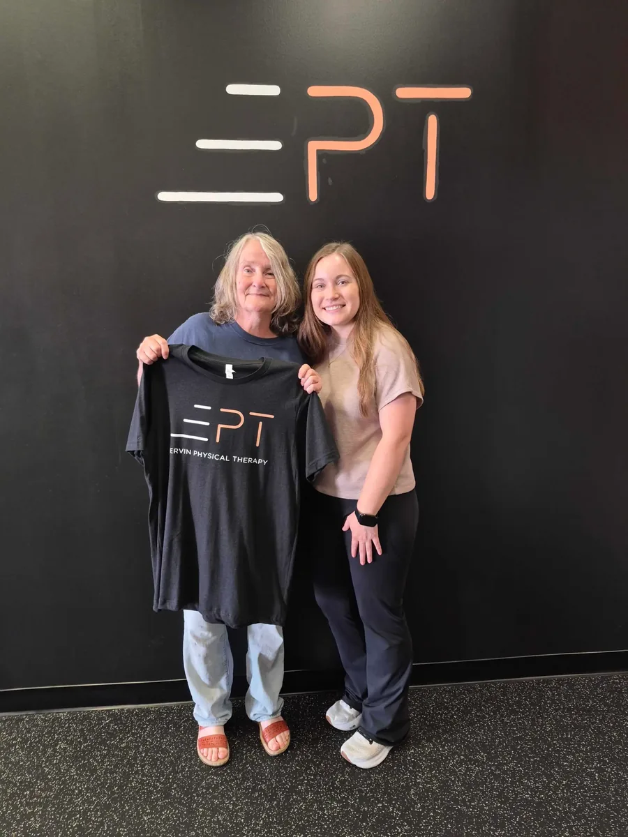 Patient holding a black EPT shirt beside their therapist in front of the EPT logo wall