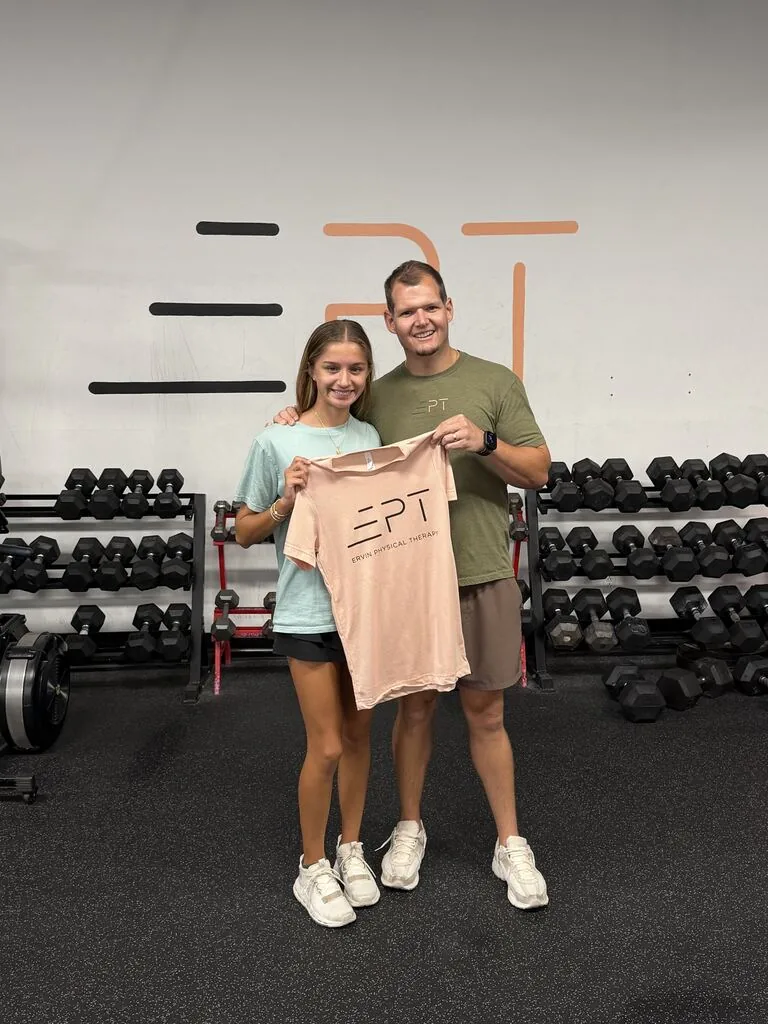 Patient holding a pink EPT shirt beside their therapist in the clinic gym