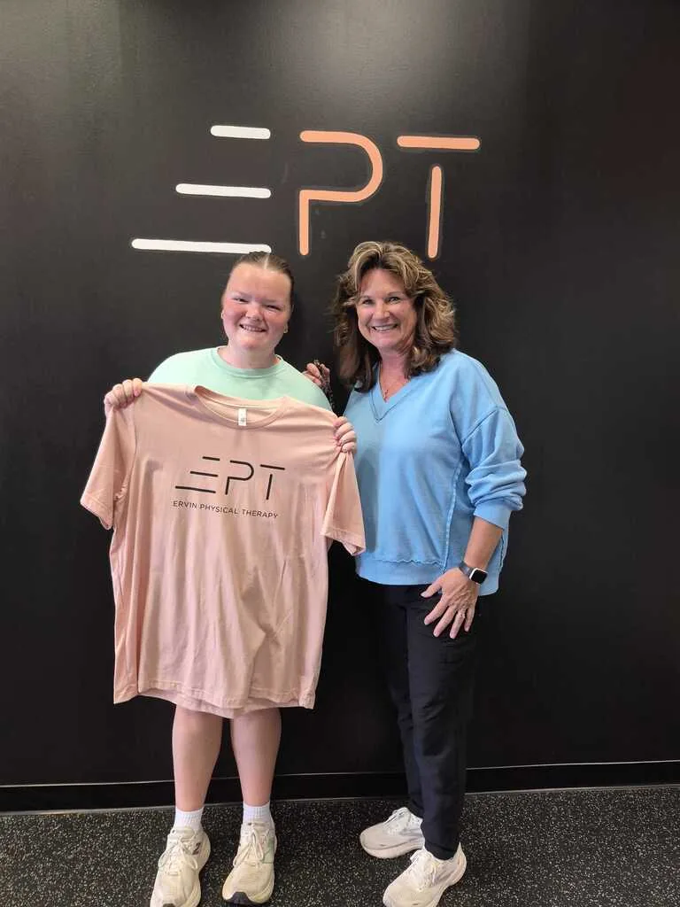 Patient holding a pink EPT shirt beside their therapist in front of the EPT logo wall
